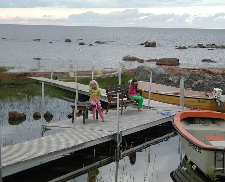Två barn sitter på en brygga vid havet med båtar och klippor i bakgrunden.