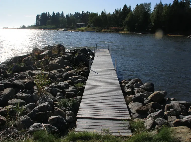 Lång brygga i trä som leder ut i vattnet, omgiven av stenar och natur, med solen som glittrar i havsytan.